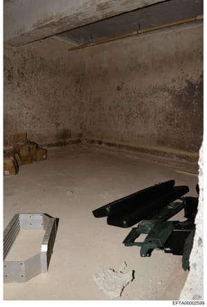 This is an evidence photograph showing an unfinished interior room with deteriorated concrete walls and floor. The space contains various items including cardboard boxes, black objects (possibly furniture or equipment), and metal/mechanical component