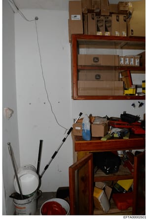 This is an evidence photograph showing a storage or utility room containing various cardboard boxes, equipment, and supplies organized on wooden shelving. The image appears to be from an property inspection or evidence documentation, featuring storag