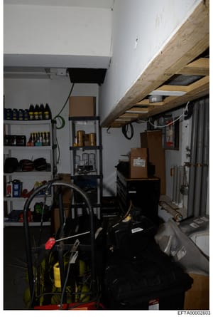 This is a photograph of a cluttered storage space or garage room containing various tools, equipment, and supplies. The image shows shelving units stocked with bottles and containers, bicycles, cardboard boxes, wooden beams overhead, electrical wirin