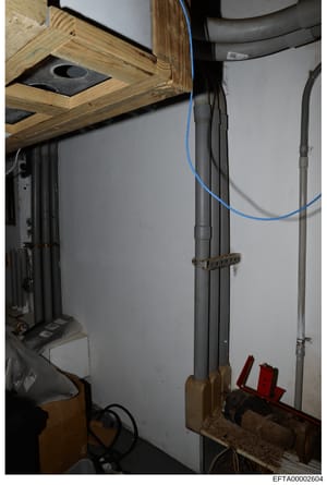 This photograph documents the interior plumbing and electrical systems of what appears to be a basement or utility room, showing PVC piping, copper conduit, blue electrical cables, and mounting hardware. The image captures infrastructure details that