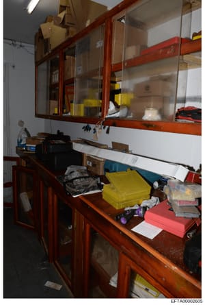 This is an evidence photograph showing an interior storage room with wooden shelving units containing various boxes, containers, and materials. The image captures organized storage with labeled items in yellow and red containers, sealed boxes on shel
