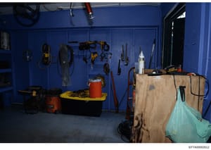 This is a photograph of a blue-walled garage or workshop space containing various tools, equipment, and storage items. The image shows organized tool storage including power tools, hand tools, extension cords, and safety equipment mounted on pegboard