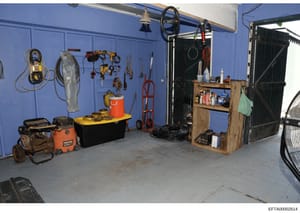 This is a photograph of an organized garage or storage space with bright blue walls. The space contains various tools, equipment, and supplies mounted on walls and stored on shelves, including power tools, chains, pulleys, buckets, a red hand truck, 