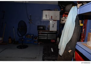 This is an evidence photograph showing the interior of a garage or workshop with blue walls, work benches, tools, and storage equipment. The image appears to be part of a property inventory or forensic documentation from the investigation.
