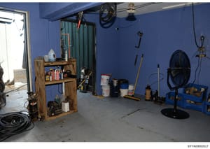 This is an evidence photograph showing the interior of a garage or workshop area painted bright blue. The space contains various tools, equipment, storage containers, and maintenance supplies organized on wooden shelving and walls. The photograph app