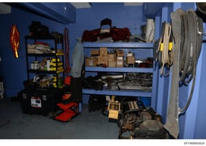 This is a photograph of a storage or utility room with bright blue walls containing various tools, equipment, and supplies. The room includes metal shelving units with cardboard boxes, automotive parts, electrical equipment, coils of wire or rope, an