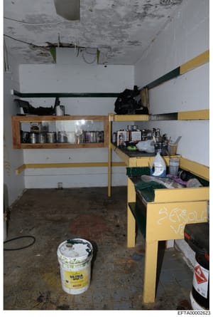 This photograph shows the interior of a basement or utility storage room, featuring deteriorated conditions with water damage visible on the ceiling, metal shelving units with various stored items, and a yellow and green storage bin system. The room 