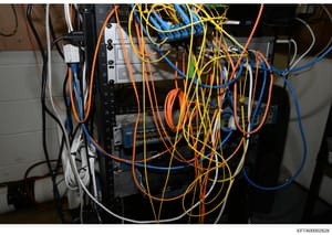 This photograph shows a close-up view of electrical or networking equipment with multiple colored cables (orange, yellow, blue, black, and white) connected throughout. The image appears to be documentation of technical infrastructure, possibly from a