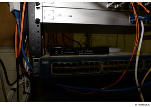 This is a photograph of network infrastructure equipment, showing a rack-mounted setup with UniFi networking equipment, various cables (orange, blue, yellow), and what appears to be server or network switching hardware. The image documents IT infrast