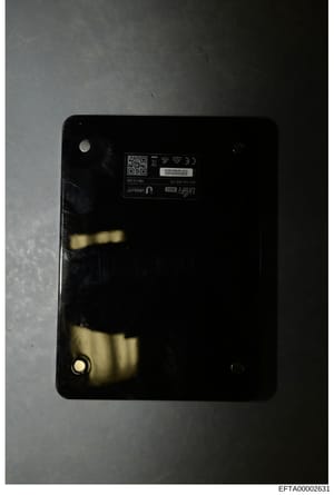 This is a photograph of the back panel of an electronic device, showing regulatory markings, a QR code, and device identification information. The image appears to be evidence documentation, with visible wear and discoloration on the black surface. T