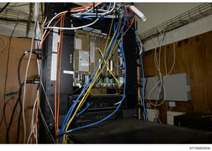 This photograph shows a complex installation of electronic equipment including servers, networking hardware, and extensive wiring with color-coded cables (orange, blue, yellow, white). The image appears to be documentation of infrastructure found dur