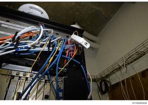 This photograph shows computer networking equipment, including routers, switches, and colored Ethernet cabling in what appears to be an interior installation at one of Epstein's properties. The equipment is mounted on a wall or panel with visible lab