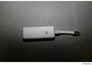 This is a photograph of a white USB device or adapter with an attached cable, documented as physical evidence in the investigation. The device is photographed against a gray concrete or stone surface. The image includes a Bates stamp indicating its e