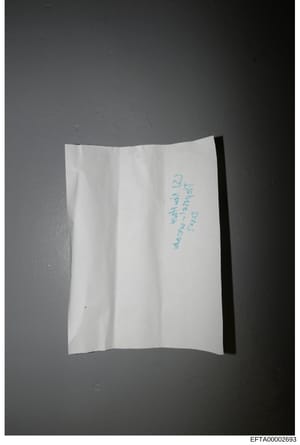 This is a photograph of a white folded piece of paper with handwritten text in blue ink. The handwriting is difficult to read clearly in the image, but appears to contain cursive writing. The document is photographed against a dark gray background. W