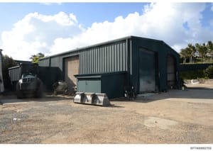 This photograph shows a large industrial metal-frame warehouse or storage building with corrugated steel siding and multiple rolling doors. The structure is situated on a sandy/gravel lot with palm trees visible in the background, suggesting a tropic