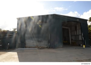 This is an evidence photograph showing a large metal-sided industrial or storage building with corrugated metal walls and a flat roof. The structure features metal gates/doors on the right side and appears to be located in a tropical or warm climate 