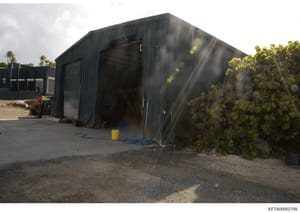 This is a photograph of a large dark metal industrial building or warehouse structure with a corrugated metal exterior. The facility appears to be located in a tropical or subtropical climate (palm trees visible in background) and shows signs of age 
