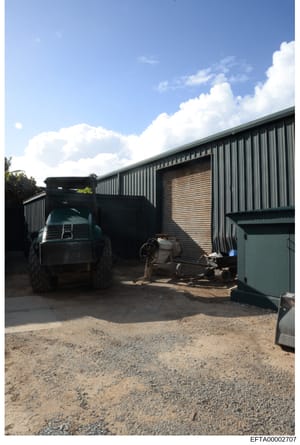 This is a photograph of an industrial or agricultural storage compound featuring a large metal-sided warehouse building with corrugated metal construction. A dark truck is parked in front of the facility, with various equipment and storage containers