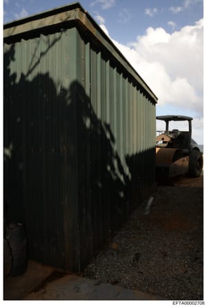 Photograph of a green corrugated metal storage building or shed photographed in daylight. A rust-colored road roller/compactor vehicle is visible to the right. The image appears to be evidence documentation of a property location, likely taken during