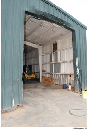 This is an evidence photograph showing the interior of a large industrial metal building with corrugated siding. The space appears to be a storage facility or garage, containing a yellow and black piece of machinery (possibly a skid steer loader or s