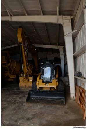 This photograph shows the interior of an industrial storage facility or equipment yard containing yellow CAT (Caterpillar) construction machinery, including an excavator and skid loader, parked under a metal-framed roof structure. The image appears t