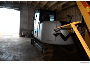 This photograph shows a large piece of heavy construction equipment (excavator) and a white trailer stored inside an industrial warehouse or hangar facility. The image appears to be documentary evidence of seized or inventoried assets, likely taken d