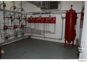 This photograph documents a commercial fire suppression system installation in what appears to be a mechanical or utility room. The image shows red pressurized tanks, control panels, piping networks, and gauges typical of an automated fire suppressio
