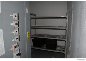 This photograph documents an electrical/server room containing a wall-mounted electrical panel with multiple circuit breakers and switches on the left side, and an open metal shelving unit on the right containing networking or server equipment. The i