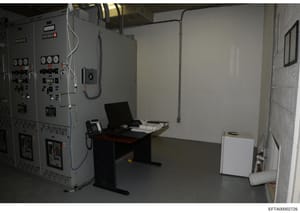 This is a photograph of an interior room containing large industrial electrical control panels and monitoring equipment. The space appears to be a utility or technical facility room, featuring grey metal equipment cabinets with various switches, gaug