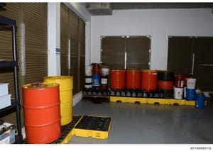 This photograph depicts an interior storage facility containing multiple large drums of various colors (red, yellow, orange, blue) and smaller containers, arranged on yellow spill containment pallets. The room features metal shelving units, pegboard 