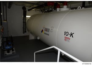 Photograph of a large white cylindrical industrial storage tank labeled '10-K' with a danger warning label, located inside a warehouse or facility. The tank appears to be a propane or fuel storage container with regulatory markings and safety signage