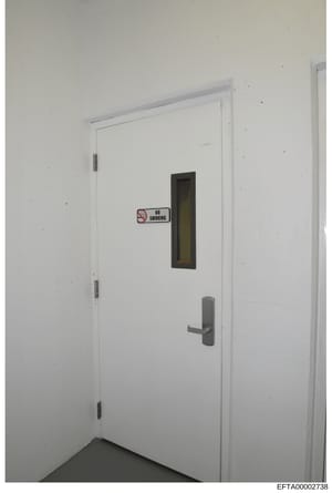 This is a photograph of an interior doorway, likely from one of Epstein's properties. The door is white with a metal handle, hinges, and a small window with dark frame. A standard 'No Smoking' sign is affixed to the door. The image appears to be evid