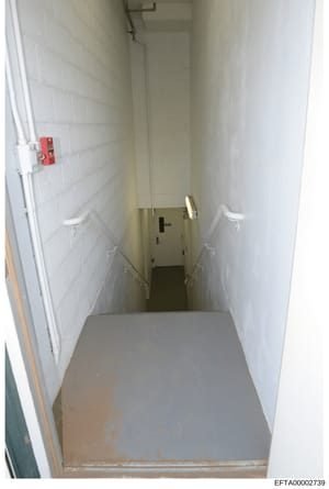 This photograph shows a narrow, institutional-style hallway or stairwell with white tile walls, concrete flooring, and a red fire alarm box mounted on the left wall. The space appears to be a basement or lower-level corridor, possibly from one of Eps
