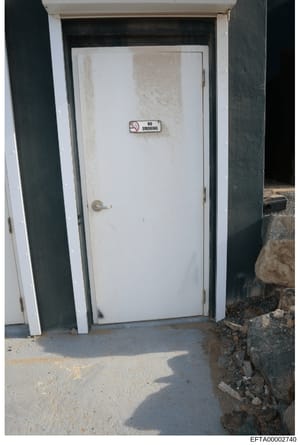 Photograph showing a white exterior door bearing a 'No Smoking' sign, located at what appears to be an outbuilding or property structure. The door shows visible wear and discoloration on its surface. This appears to be an evidence photo documenting p
