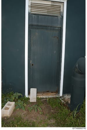 This is a photograph of an exterior door to a building, showing a weathered teal/dark blue painted door with visible rust stains and deterioration. The door features a transom window above it and is set within a white-trimmed frame. The ground area s
