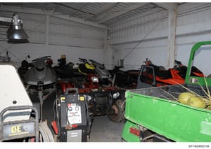 This photograph shows the interior of a large metal warehouse or storage facility containing multiple personal watercraft (jet skis), ATVs, power washing equipment, and other recreational vehicles and tools. The image appears to be documentation of a