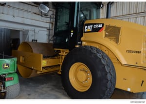 This photograph shows heavy construction machinery stored in an industrial warehouse, including a Caterpillar CS54B compactor roller and other equipment. The image appears to be a property inventory or asset documentation photo, possibly related to e