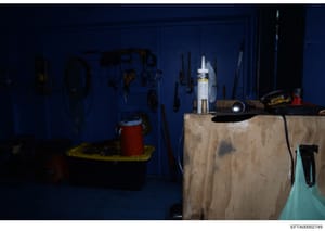 This is an evidence photograph showing a storage area or workshop space with bright blue walls. Various tools and equipment are visible, including hand tools hung on the wall, a red bucket or container on the left, and work surfaces with construction