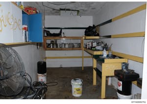 This photograph documents an interior storage or basement room containing yellow metal shelving units, various maintenance and chemical supplies, tools, and equipment. The space appears to be part of a property inventory or evidence documentation, sh