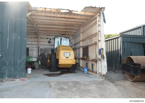 Photograph of the interior of an industrial or construction equipment storage facility showing a yellow Caterpillar (CAT) road roller/compactor parked inside a large metal-framed warehouse structure. The image documents equipment and facility conditi