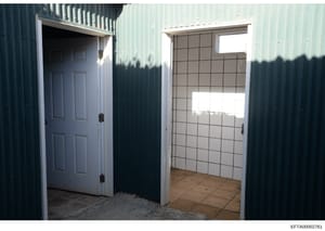 This photograph shows the exterior and interior entrance of a metal-sided structure, featuring a white six-panel door on the left and a tiled interior room visible through an open doorway on the right. The image appears to be evidence documentation f