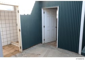 This photograph shows an interior or semi-enclosed space featuring white paneled doors and green corrugated metal wall panels. The structure appears to be a utility or storage area with tiled flooring visible on the left side and concrete/gravel floo