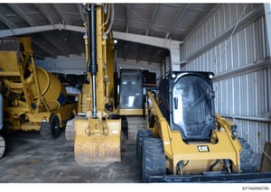This photograph shows the interior of a large industrial storage facility or warehouse containing heavy construction equipment. The image depicts multiple yellow Caterpillar (CAT) branded pieces of equipment including excavators, a concrete mixer tru