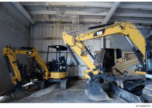 This is a photograph of an industrial warehouse or storage facility containing multiple pieces of heavy construction equipment, primarily yellow Caterpillar (CAT) excavators. The image shows at least three excavators of varying sizes stored in a meta