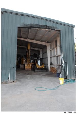 This photograph shows the interior of a large metal-frame warehouse or industrial storage building with corrugated metal siding. The space contains heavy machinery including what appears to be construction or industrial equipment (yellow excavator or