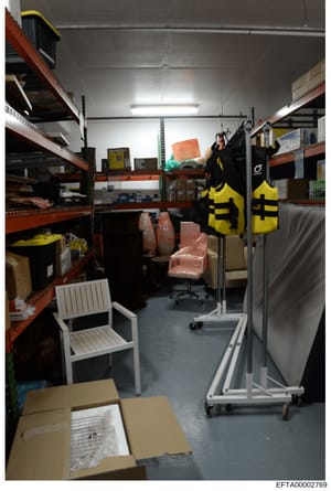 This photograph documents the interior of a storage or warehouse space containing various items including furniture, recreational equipment, and boxed goods. The space features industrial orange shelving units, yellow and black life vests or safety e