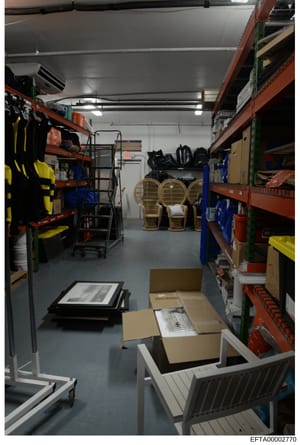 This photograph shows the interior of a warehouse or storage facility containing various items of furniture, sports equipment, and boxed goods on industrial shelving. The image appears to be evidence documentation from the investigation, showing seiz