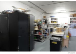 This is an evidence photograph showing an interior storage or utility room containing various equipment, shelving units, boxes, and supplies. The space appears to be well-organized with labeled storage containers (including one marked '2'), metal she