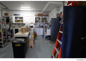 This photograph shows the interior of a storage room, workshop, or utility space containing various tools, equipment, and supplies organized on shelves and work surfaces. The image appears to be evidence documentation from the investigation, though t