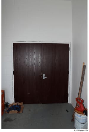 This is a photograph of an interior space showing a large dark wooden double-door entry with a metal handle, set in a white cinder block or painted brick wall. The room appears to be under construction or renovation, with various tools and materials 